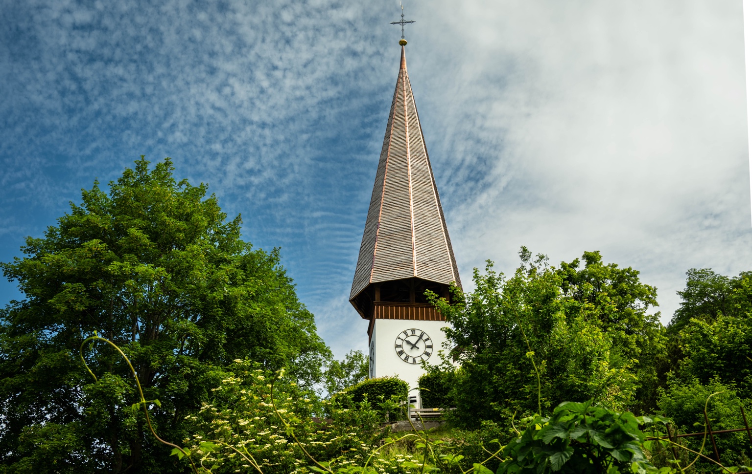 Kirchturm zwischen grünen Bäumen unter lebhaftem Wolkenhimmel